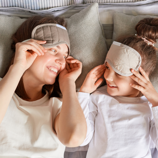 Mom and daughter lying in bed together. They are wearing face masks on their foreheads and smiling at eachother.